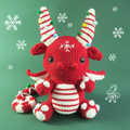 Red and white crocheted dragon toy with candy cane horns on a green background with snowflakes.