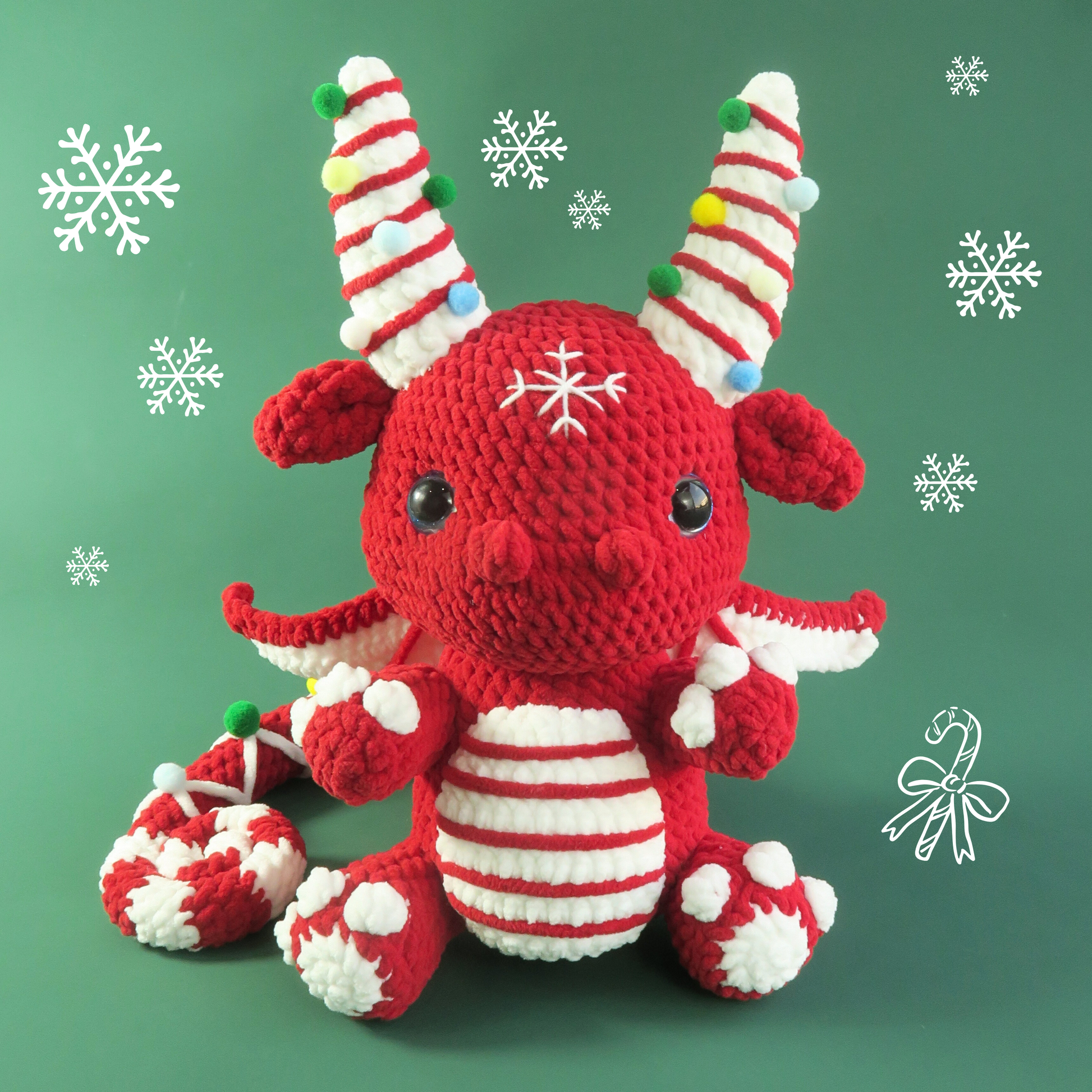 Red and white crocheted dragon toy with candy cane horns on a green background with snowflakes.
