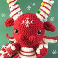 Crocheted dragon toy with candy cane designs on a green background with snowflakes.