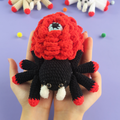 Hand holding a Eyes Rose Spider Crochet Pattern against a purple background with scattered yarn balls.