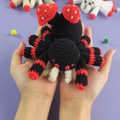 Hand holding a mushroom crochet spider toy against a purple background.
